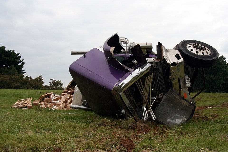 Purple semi truck that appears to have been in a rollover accident. Cargo or freight of boxes spilled from the back of the semi. The truck is on grass and dirt, possibly a median of a road.