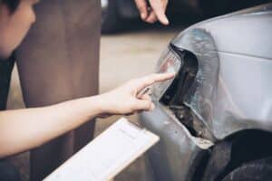 Person inspecting front-end vehicle damage after a collision, pointing to a damaged headlight and dented bumper while documenting repairs, representing Lyft accident claims, vehicle damage assessment, and rideshare injury cases in San Diego.